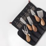 A full set of six Esie Houzie stainless steel cheese knives with acacia wood handles, neatly arranged in their storage bag.